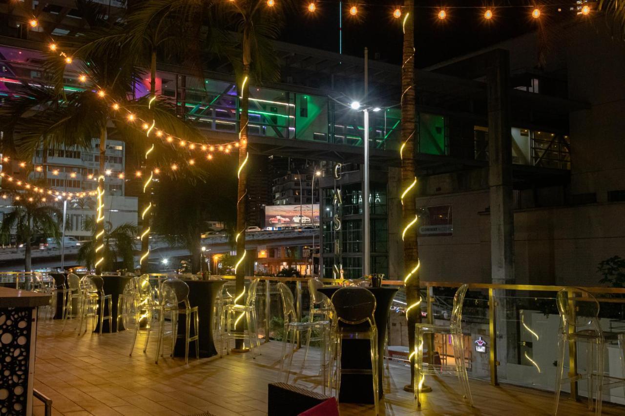 Decapolis Hotel Panama City Exterior photo The photo shows an outdoor seating area, likely at a restaurant or bar. It features several tall tables with transparent chairs around them. The space is illuminated with string lights that create a warm and inviting atmosphere. In the background, th