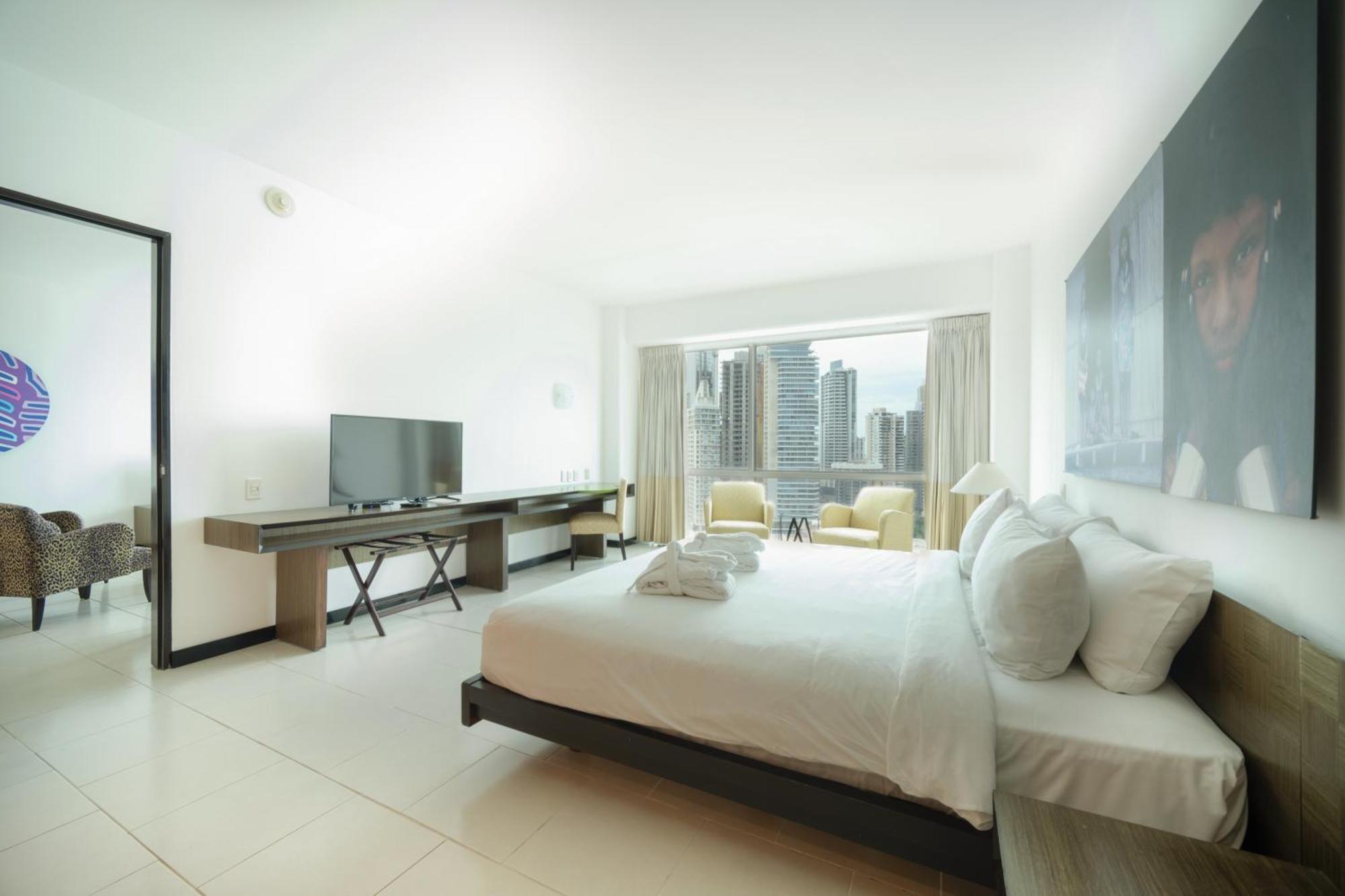 Decapolis Hotel Panama City Exterior photo The photo depicts a modern hotel room featuring a large bed with white linens and several pillows. There is a television mounted on the wall opposite the bed, and a desk with a chair beside it. Large windows provide a view of a city skyline, with two