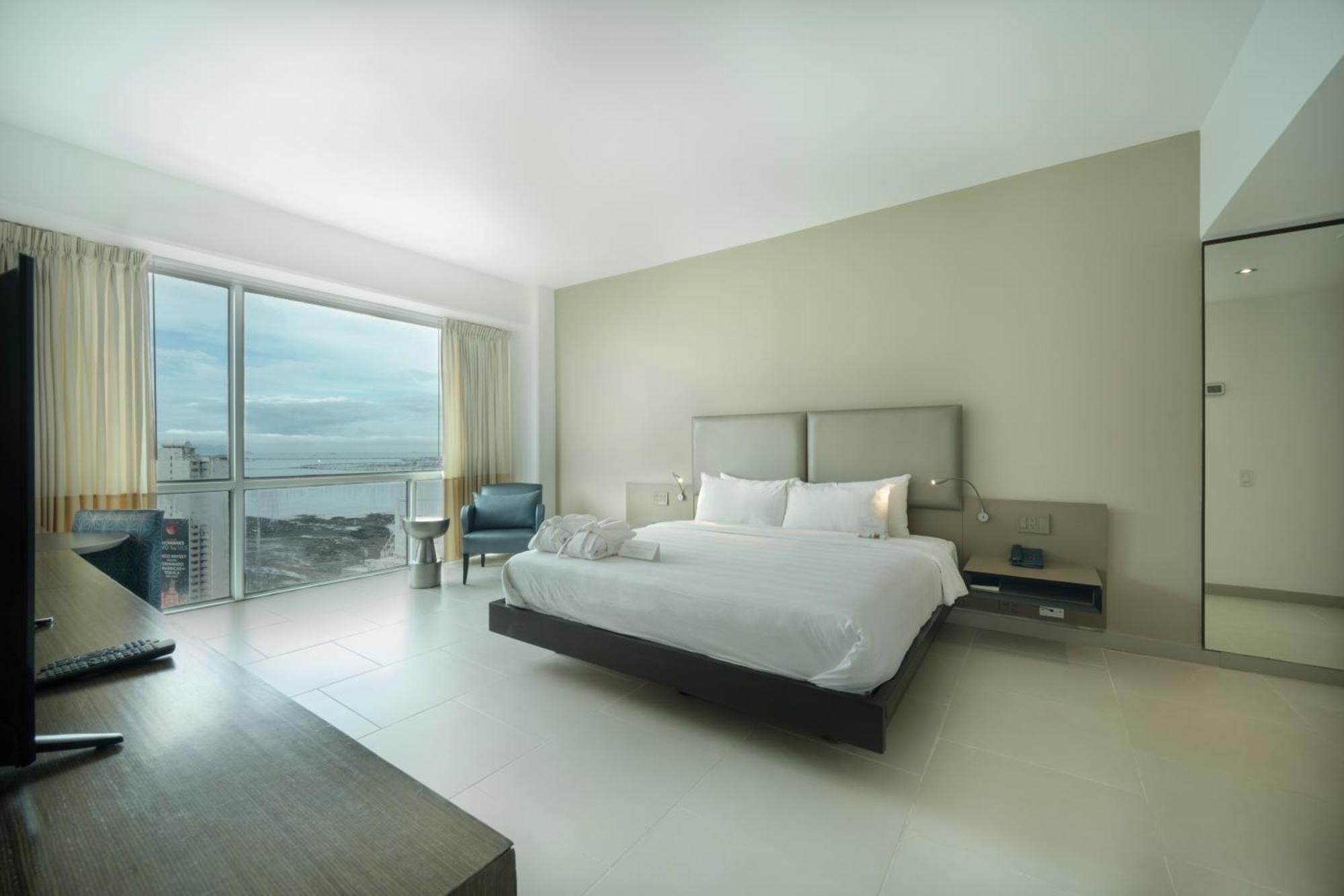 Decapolis Hotel Panama City Exterior photo The photo shows a modern and minimalist hotel room. It features a large bed with white bedding, complemented by neutral-colored walls. There is a sleek wooden desk and a chair in the corner, along with a small seating area by a window that looks out 