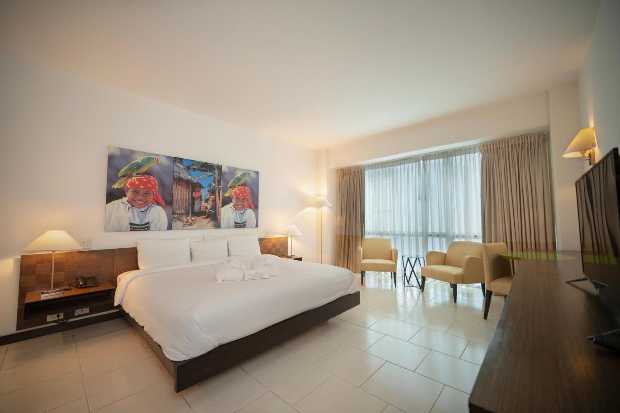 Decapolis Hotel Panama City Exterior photo The photo depicts a hotel room featuring a large bed with white linens and two pillows. In the background, there are two light beige armchairs next to a window that allows natural light to flood the space, complemented by sheer curtains. On one side 