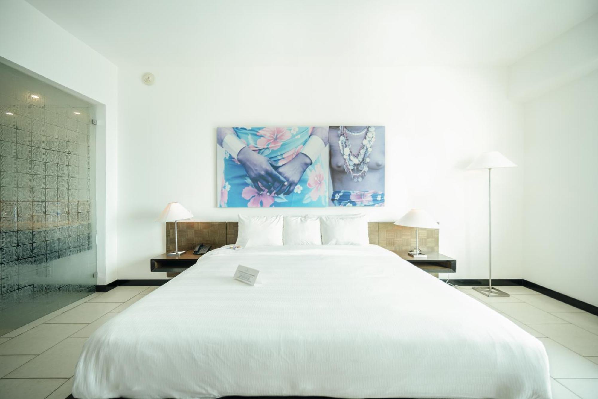 Decapolis Hotel Panama City Exterior photo The photo shows a modern bedroom with a clean and minimalist design. The bed is large with a white comforter and several pillows. On the wall above the bed, there are two framed art pieces featuring floral and jewelry motifs, adding a touch of color 
