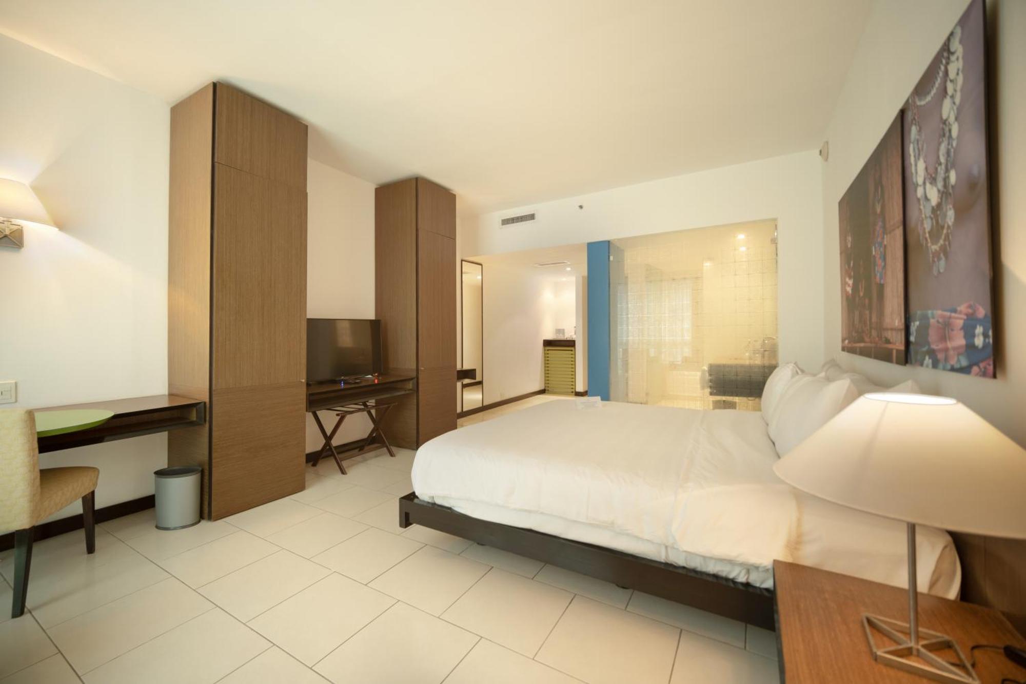 Decapolis Hotel Panama City Exterior photo The photo shows a modern hotel room. It features a large bed with white bedding, a wooden headboard, and bedside tables with lamps. There is a wooden wardrobe or storage unit on one side, along with a desk and a chair. A flat-screen TV is mounted on 