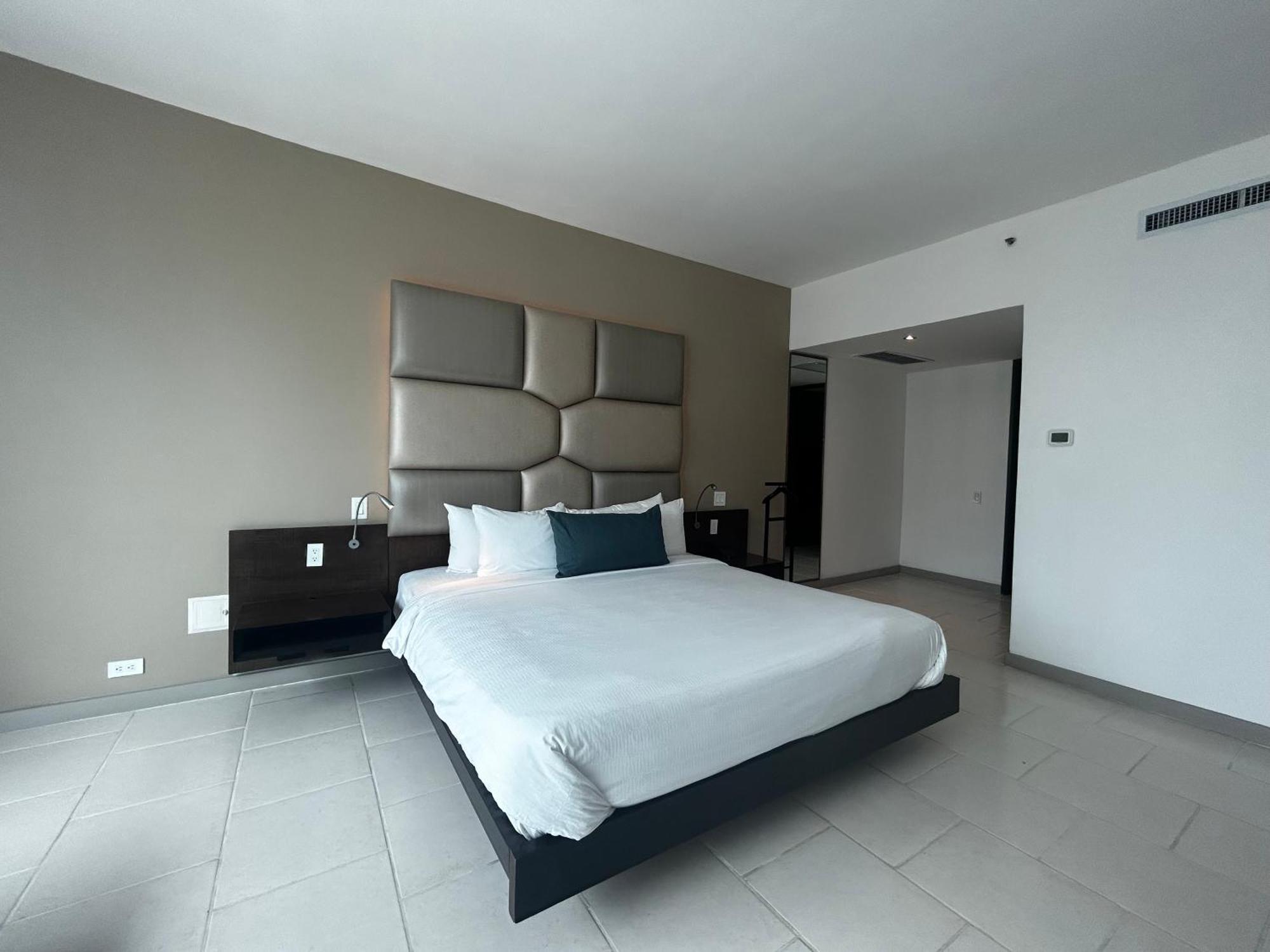 Decapolis Hotel Panama City Exterior photo The photo shows a modern hotel room featuring a spacious, minimalist design. In the center, there is a large bed with a white duvet and pillows, positioned against a light gray accent wall that has a padded headboard. To the side, there is a simple n