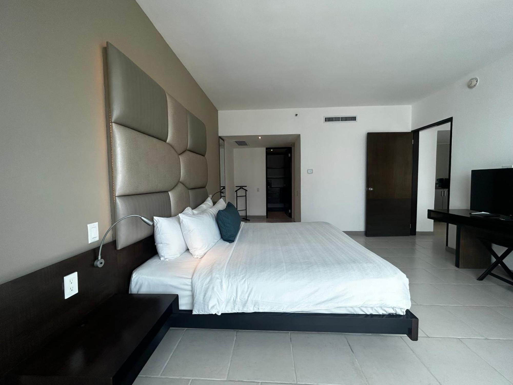 Decapolis Hotel Panama City Exterior photo The photo shows a modern hotel room. It features a large bed with a white comforter and several pillows, including a couple of blue accent pillows. The headboard of the bed is upholstered in a light gray material. Next to the bed, there is a dark woo