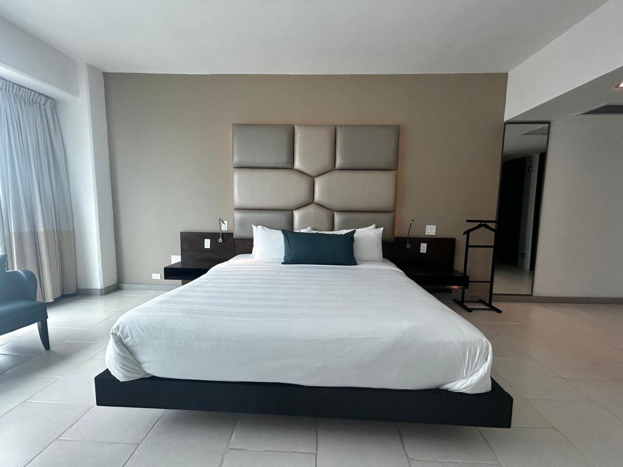 Decapolis Hotel Panama City Exterior photo The photo shows a modern and minimalist hotel room. In the center, there is a large bed with a simple white bedspread and a dark accent pillow. The headboard is composed of light-colored padded panels. On either side of the bed, there are dark wooden