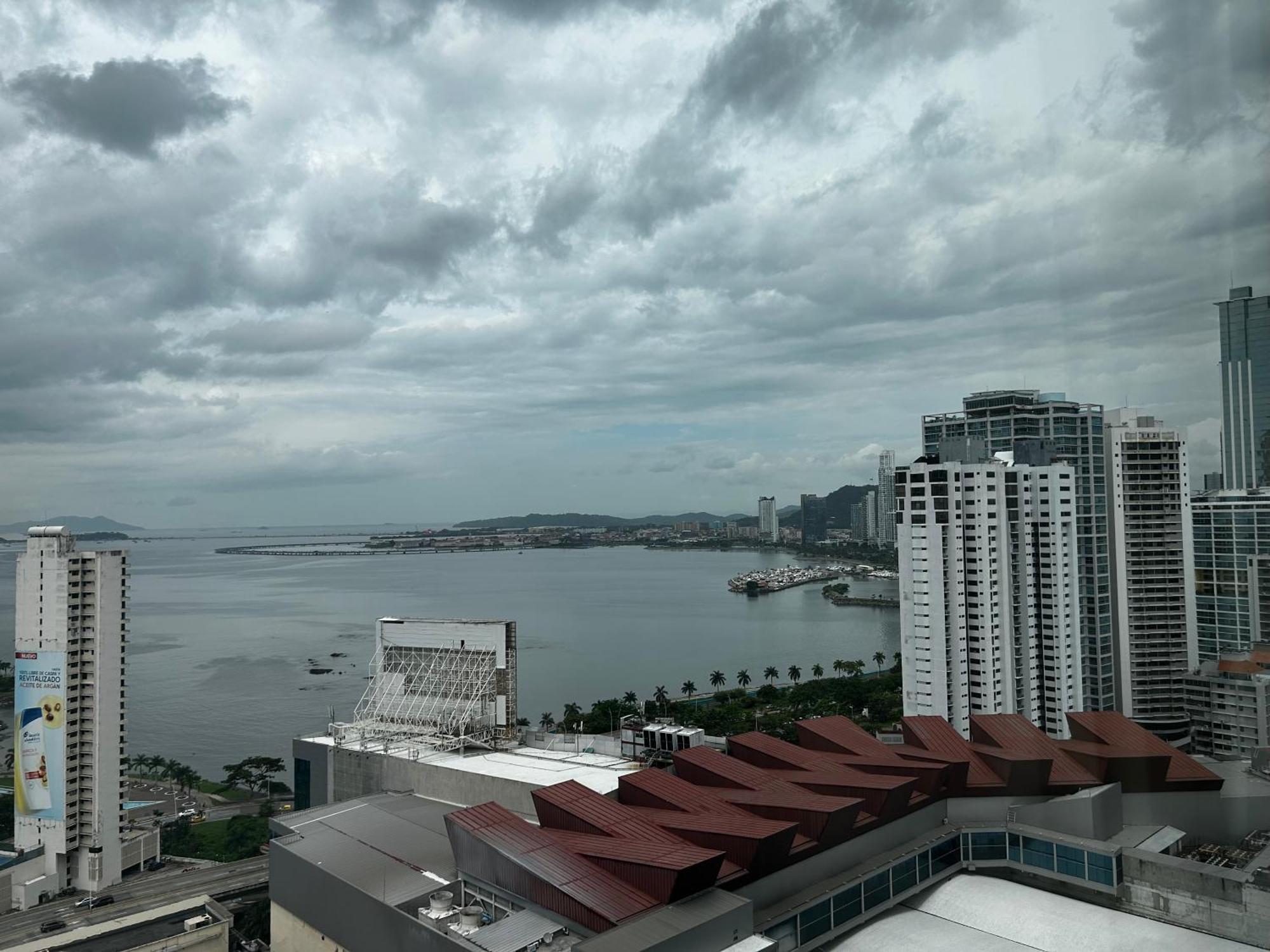 Decapolis Hotel Panama City Exterior photo The photo depicts a coastal cityscape under a cloudy sky. In the foreground, there are several modern buildings, including some with distinctive architectural features, like a red-tiled roof. In the background, a bay or harbor can be seen, with water