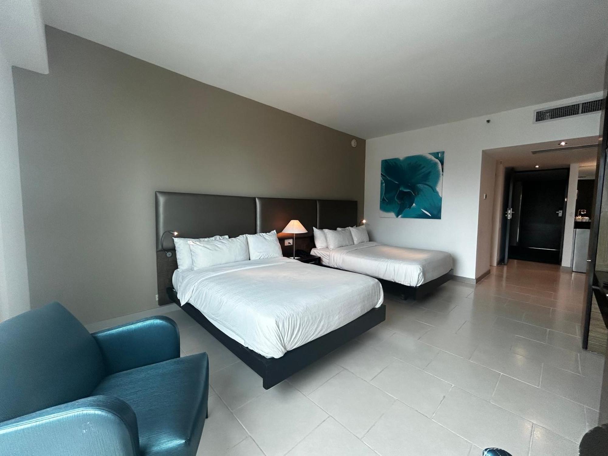 Decapolis Hotel Panama City Exterior photo The photo shows a spacious hotel room featuring two queen-sized beds, each with white bedding. There is a blue armchair in one corner, adding a pop of color to the neutral-toned decor. A large piece of artwork with a blue flower is displayed on the w