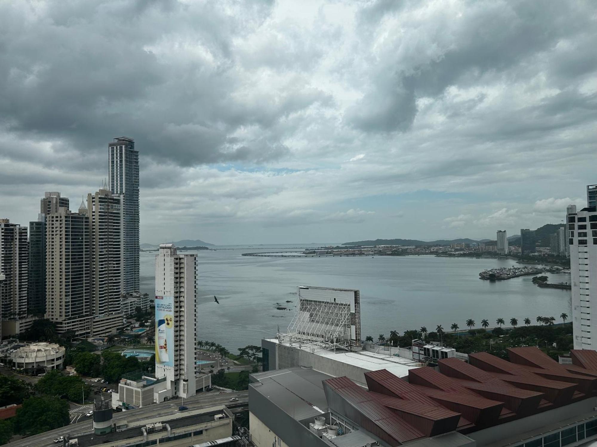Decapolis Hotel Panama City Exterior photo The photo shows a coastal cityscape featuring tall buildings and skyscrapers lining the waterfront. In the foreground, there are modern structures with distinctive architectural designs. The scene is dominated by a cloudy sky, suggesting an overcast 