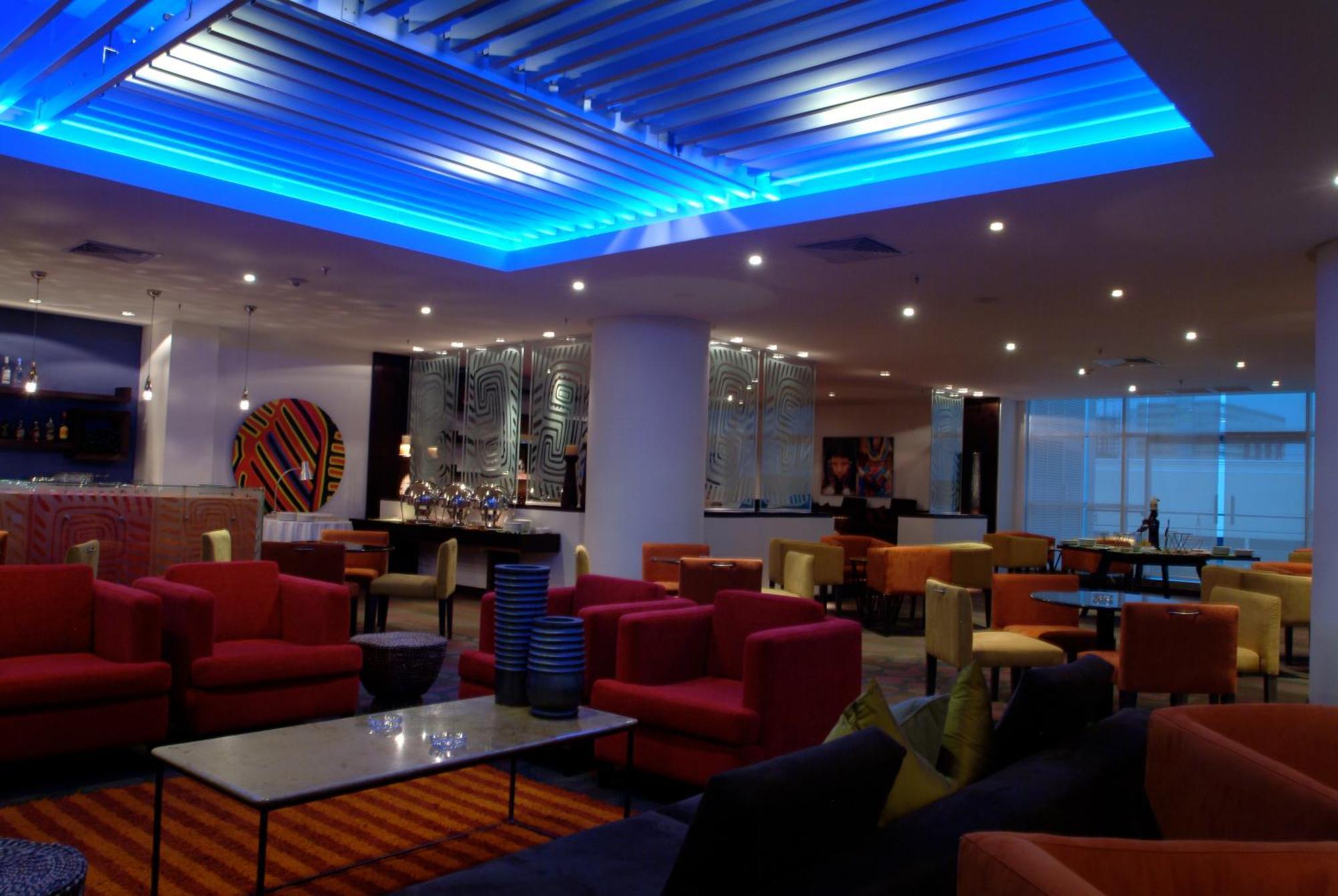 Decapolis Hotel Panama City Restaurant photo The photo shows a modern lounge or lobby area with various seating arrangements. The furniture includes colorful chairs and sofas, predominantly in shades of red, pink, and yellow, arranged around glass-topped tables. The ceiling features a blue illu